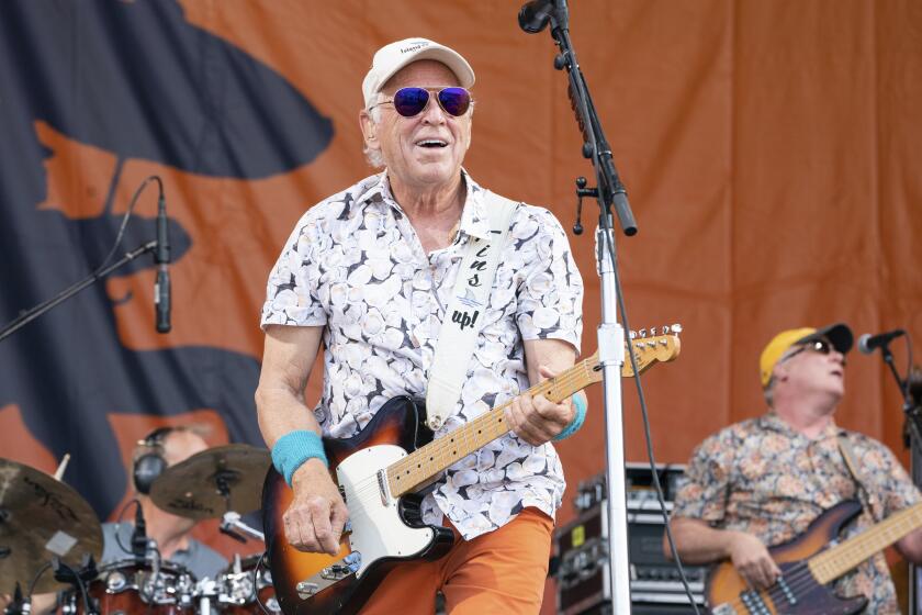 Jimmy Buffett performs while wearing flowered shirt, cap and sunglasses and is strumming electric guitar on stage. 