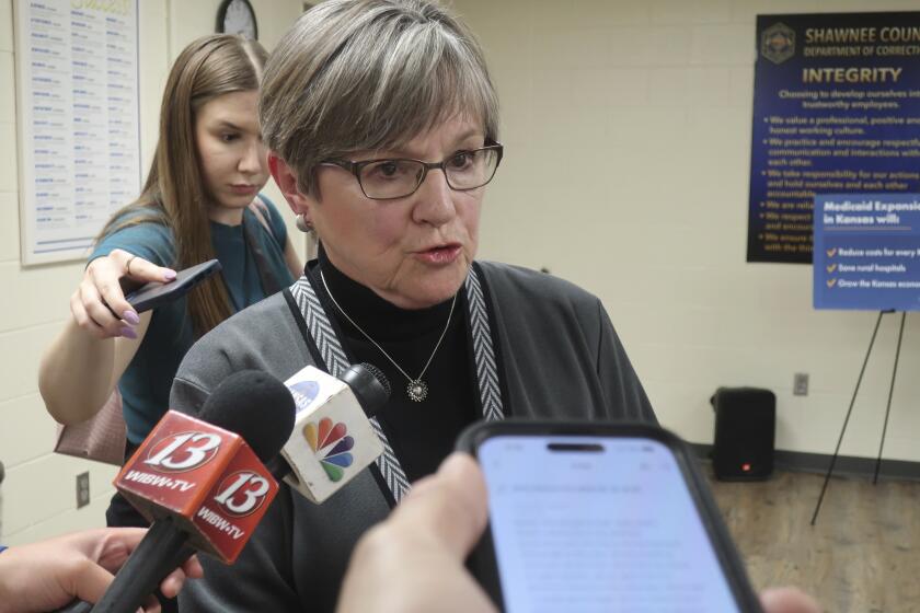FILE - Kansas Gov. Laura Kelly answers questions from reporters following an event at the Shawnee County, Kan., jail, Tuesday, April 16, 2024, in Topeka, Kan., as her communications coordinator, Grace Hoge, watches behind her. Kelly on Friday, April 29, 2024, vetoed proposed tax breaks for anti-abortion counseling centers while allowing restrictions on college diversity initiatives approved by the Republican-controlled Legislature to become law without her signature(AP Photo/John Hanna, File)