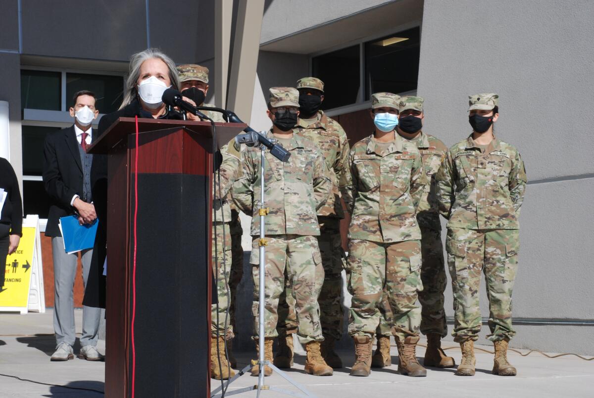 National Guard troops in uniform stand behind a woman speaking at a lectern