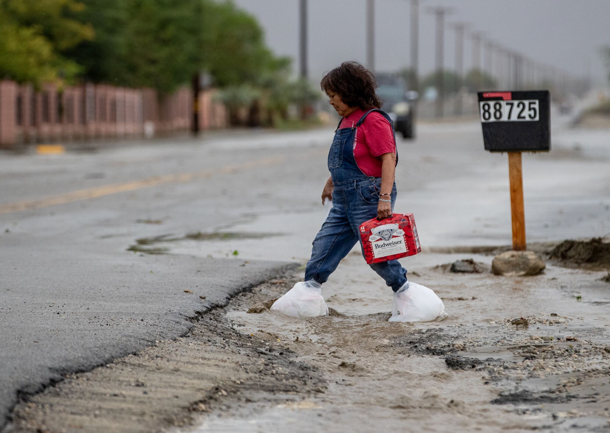 A person in red shirt and blue overalls, her feet wrapped in white plastic, approaches a street while carrying a red package 