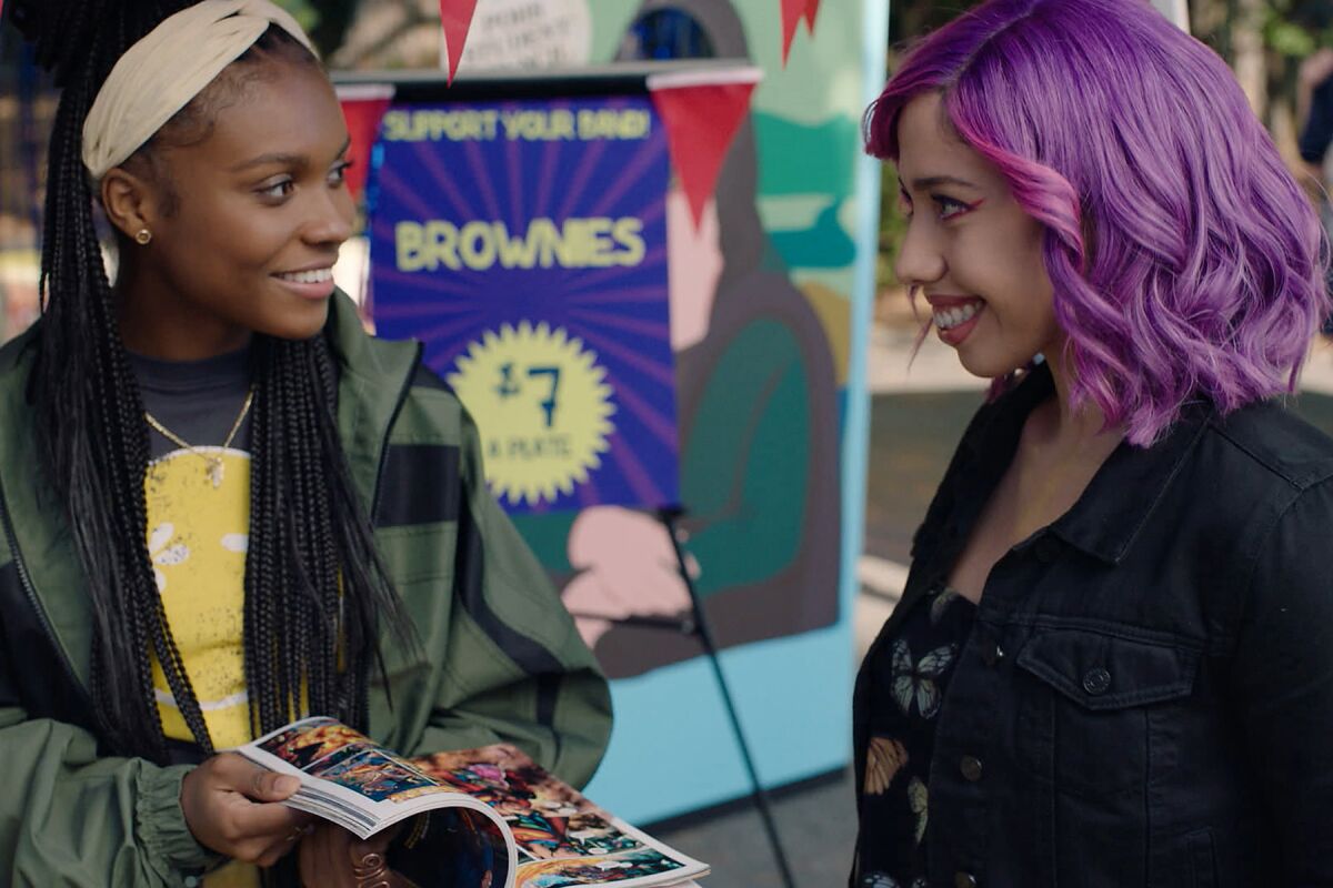 A woman with braids talks about a comic book with a woman with purple hair.