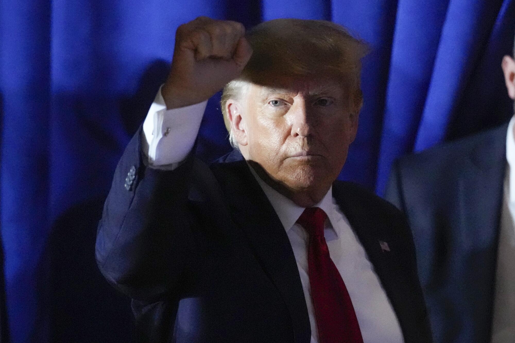 In a head-and-shoulders frame Donald Trump is lighted by a shaft of light across his face while he rises his right fist