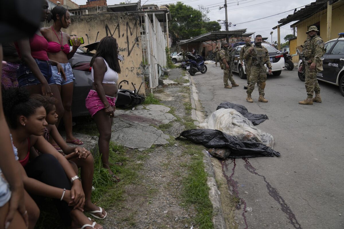 Women watch as military police patrol by the covered body of a person who was killed during a police operation against alleged drug traffickers in the Vila Cruzeiro favela of Rio de Janeiro, Brazil, Friday, Feb. 11, 2022. The operation left at least eight people dead. (AP Photo/Silvia Izquierdo)