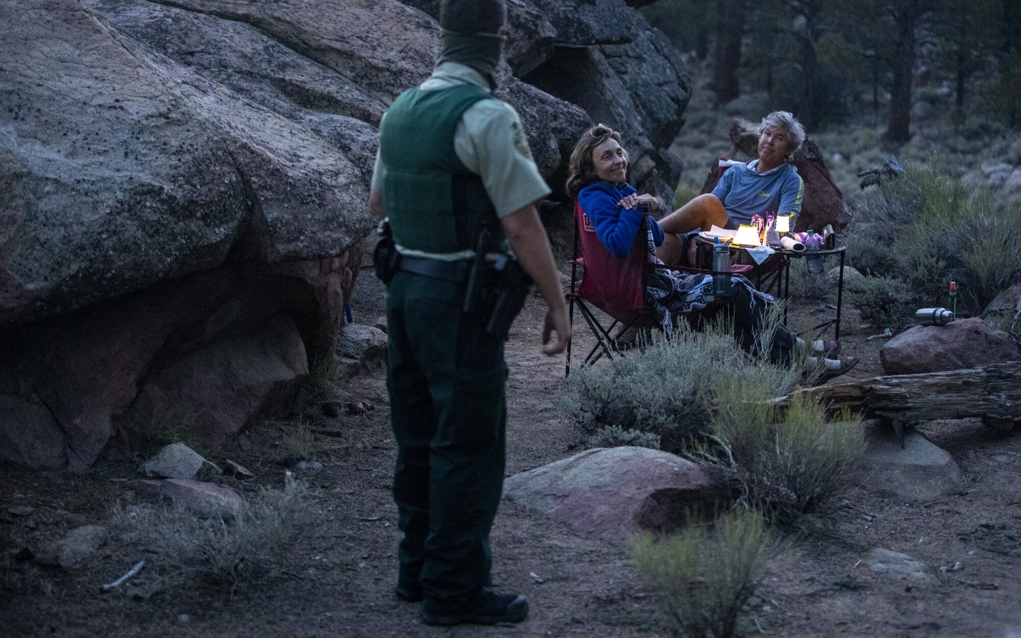 An armed ranger speaks to a couple seated at a small table in a campsite amid granite boulders and brush