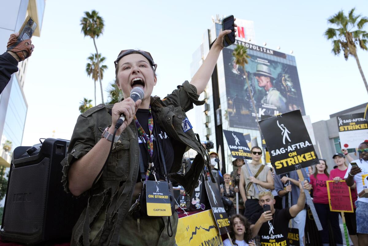 a woman in a green jacket with a microphone yells as people hold SAG-AFTRA signs
