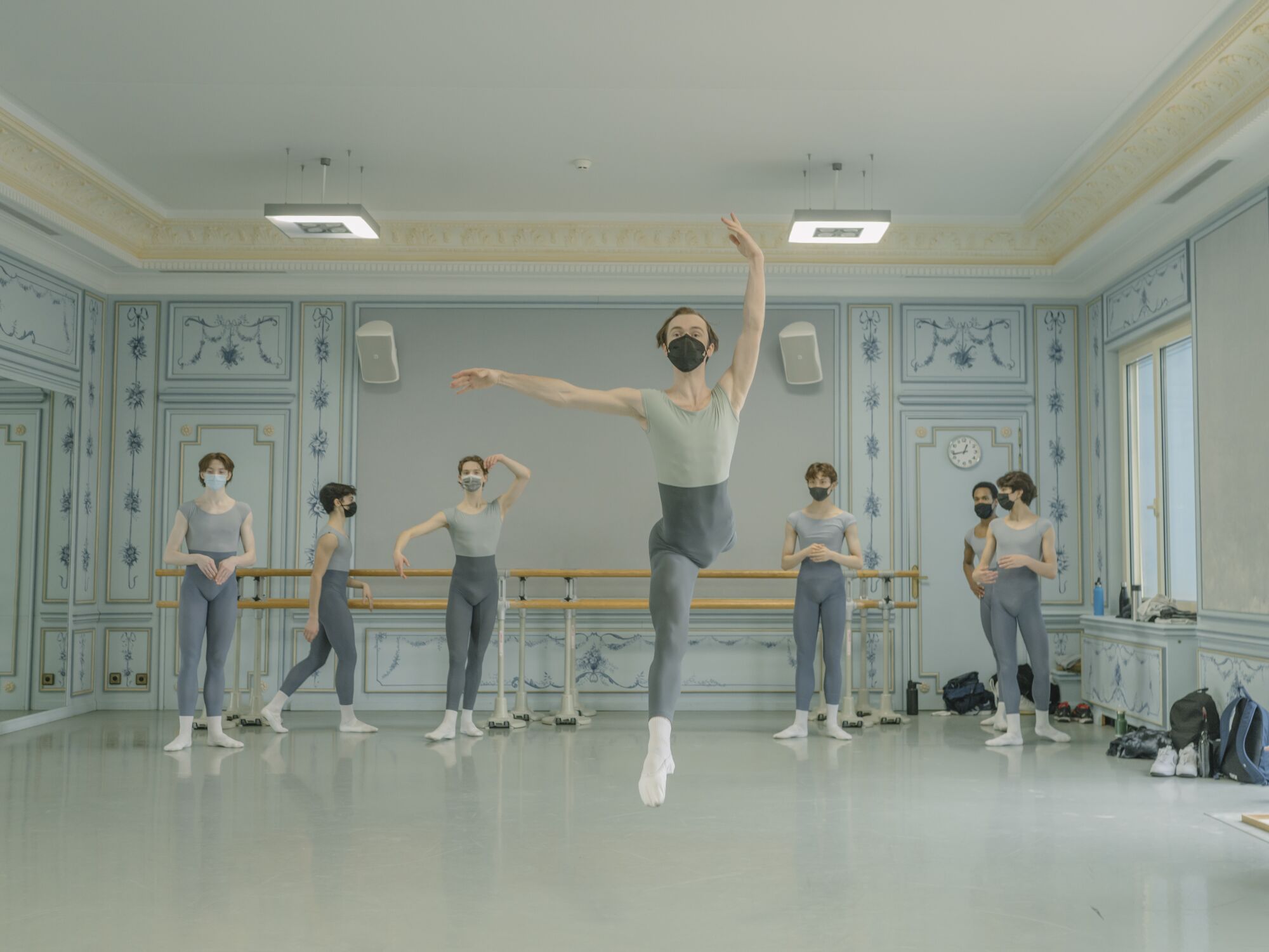 A boy leaps in a ballet studio with other boys at the bar behind him  