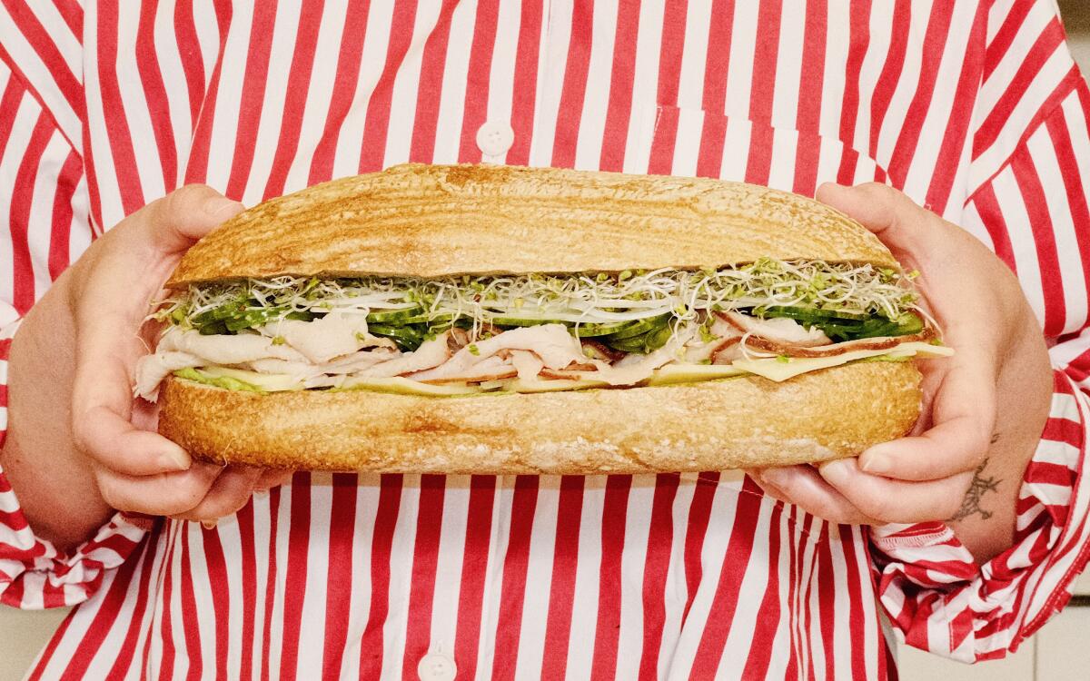 Two hands hold a whole turkey sandwich with green spread against a red-and-white striped shirt