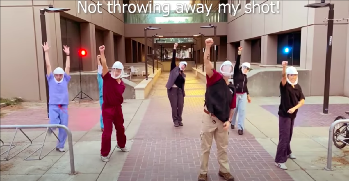 Seven healthcare workers raise their fists in a screenshot of a video with the caption "Not throwing away my shot!"
