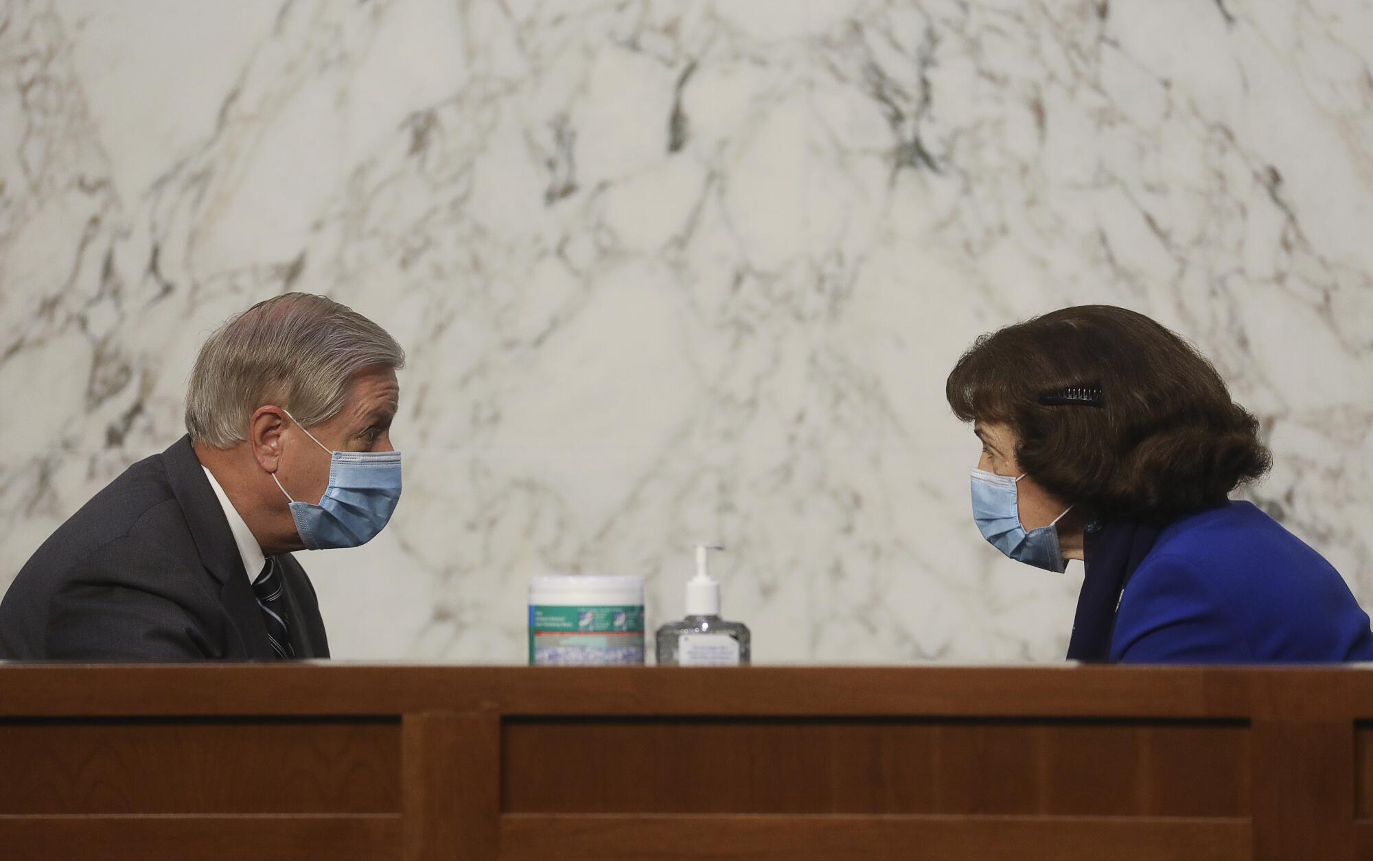 With wipes and hand sanitizer between them, Lindsey Graham and Dianne Feinstein, wearing masks, chat.