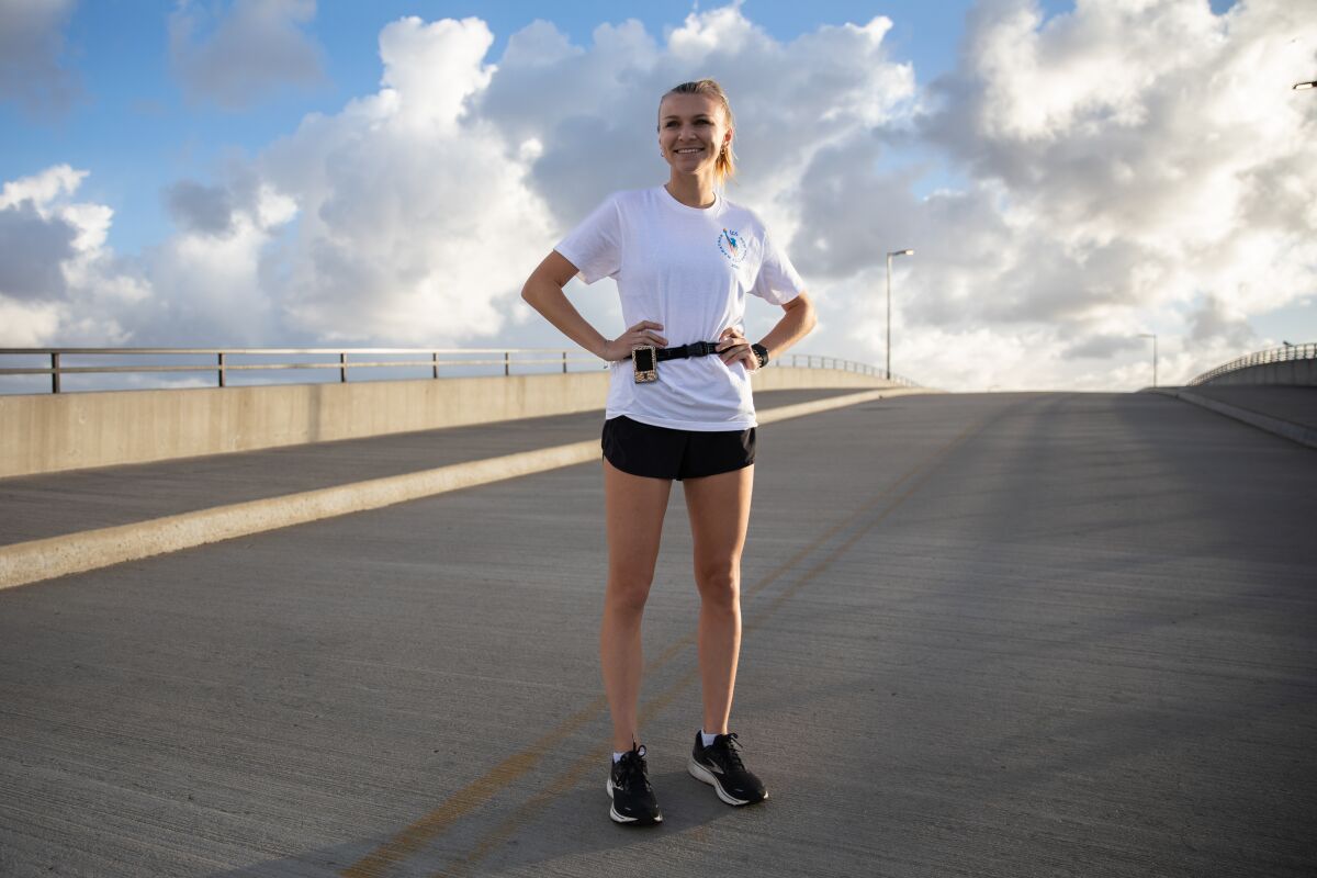 A woman wearing running shoes and a glucose monitor stands on a road bridge, smiling, against a sunny, cloud-dappled sky.