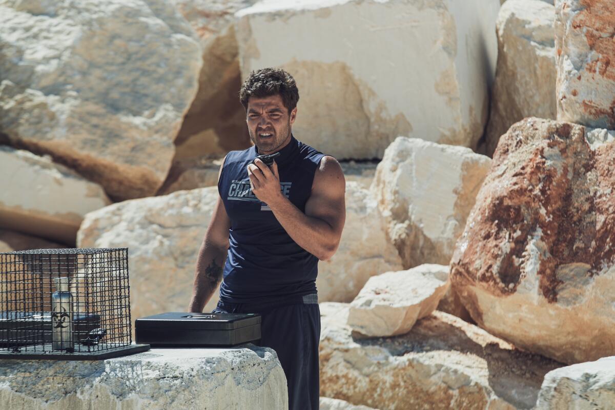 A man in black stands before a pile of rocks, surveying a briefcase and a bottle containing a toxic substance in a cage.