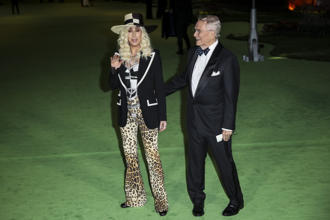 A woman in a black-and-white hat, jacket and cheetah-print pants and a man in a black suit posing on a green carpet