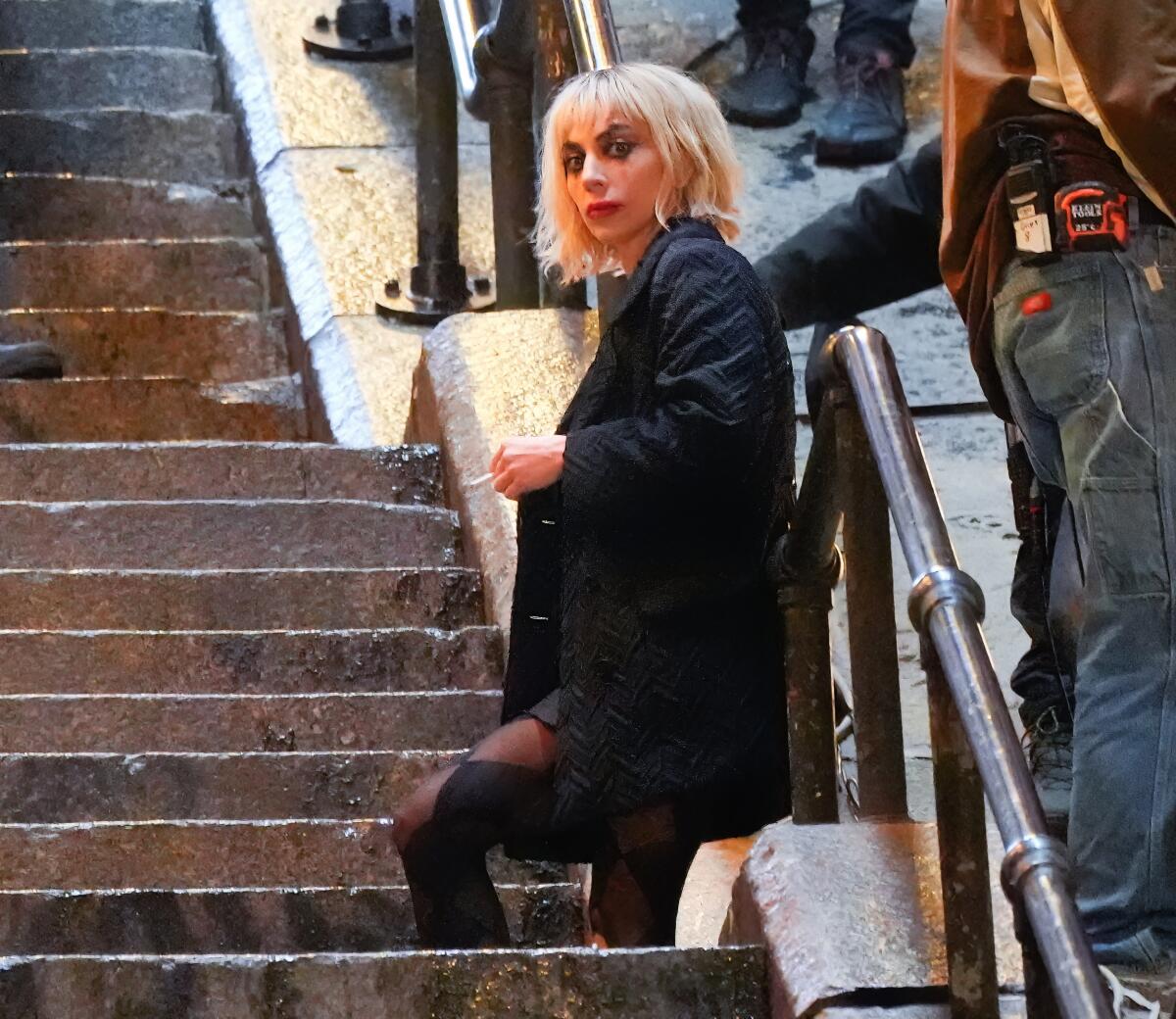 Lady Gaga leans against a railing on a staircase in character and makeup as Harley Quinn