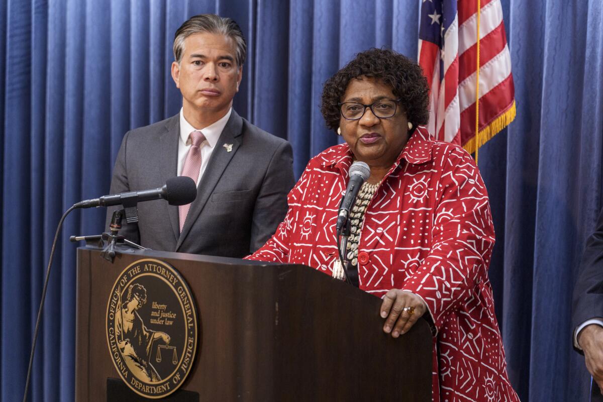 A man in a gray suit, left, and a woman wearing glasses and a red printed jacket stand at a lectern with mics