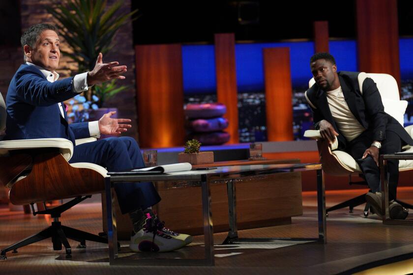 Mark Cuban and Kevin Hart sitting in chairs in a studio. Mark's wearing a suit and holding out his right hand in front of him