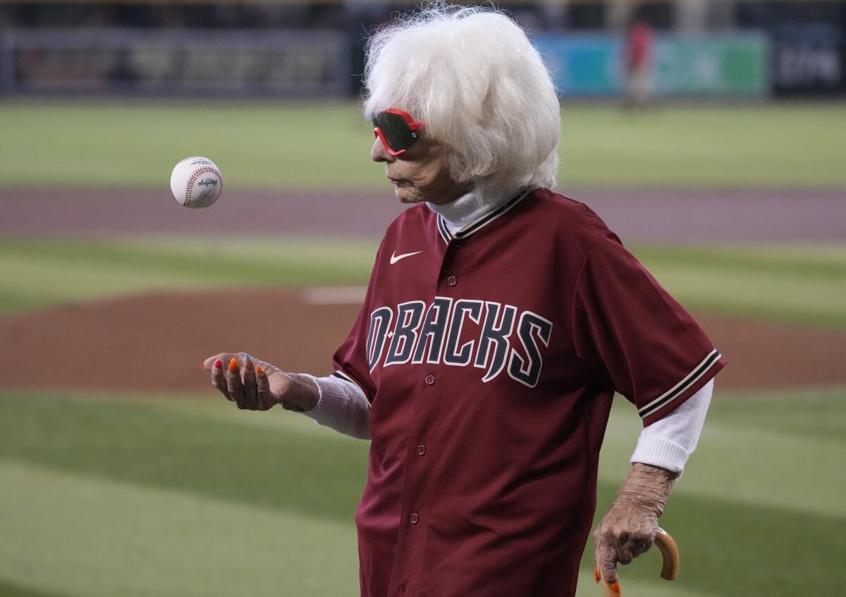 On a baseball field, an elderly woman in a D-Backs jersey and using a cane tosses a baseball in the air