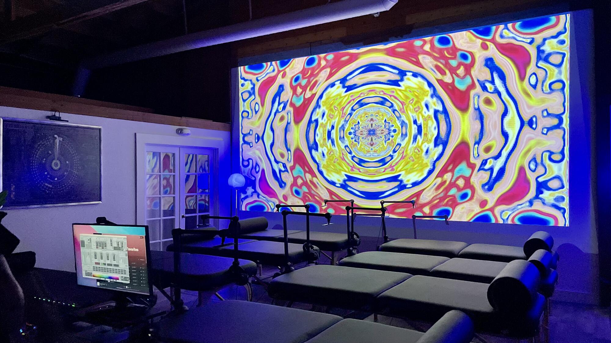 A dimly lit room with a cluster of massage tables in the center and trippy, abstract graphics on a big screen.
