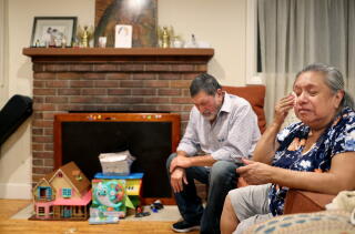 A woman wipes away tears as a man looks downward from their seats near a brick fireplace, children's toys on the hearth.