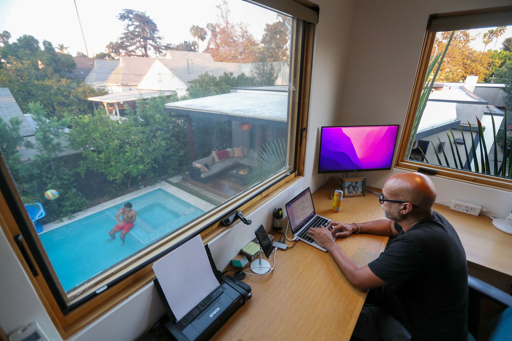 A man works at a desk next to a window that overlooks the backyard swimming pool.
