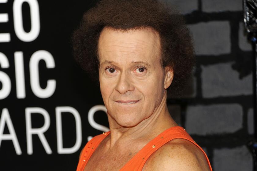 Richard Simmons in an orange exercise tank top with sparkles looks at the camera on a red carpet