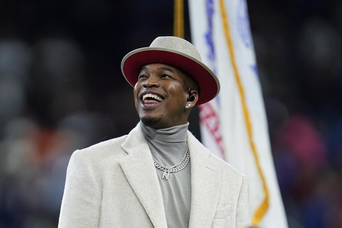Ne-Yo smiles in a light-colored suit and hat and a gray turtleneck