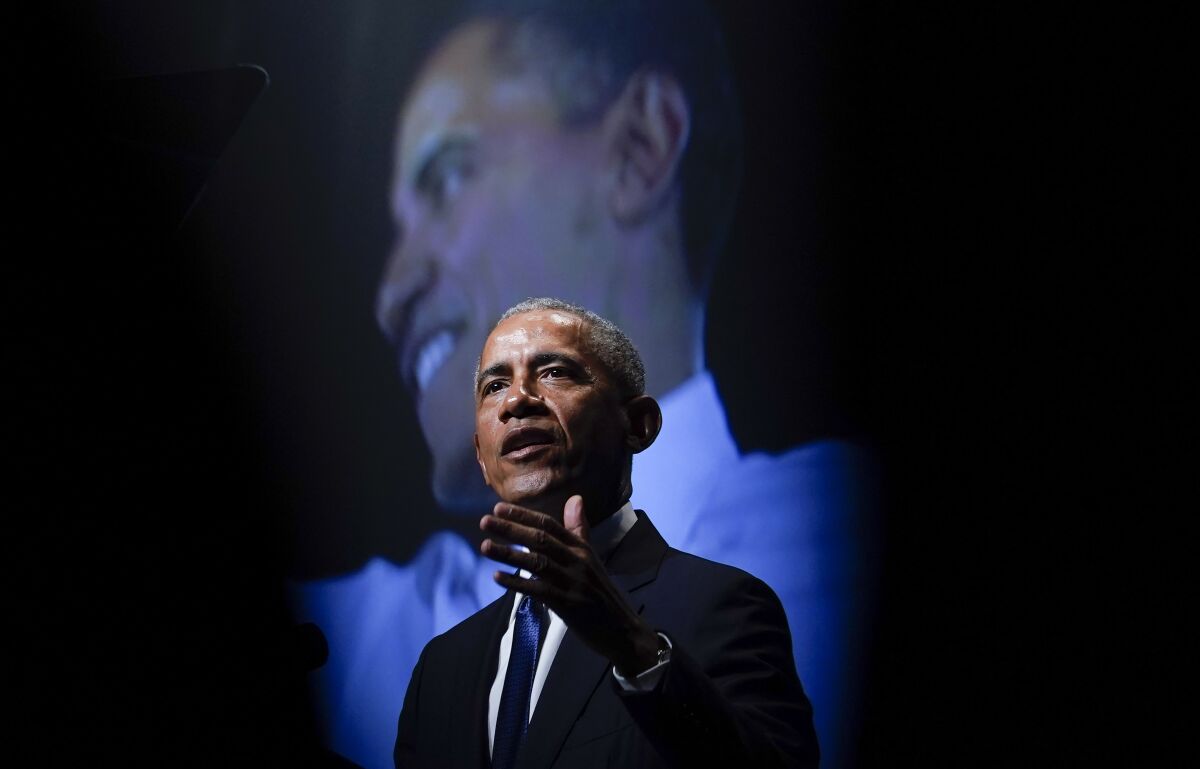 Former President Obama speaks in front of a screen with his image on it.
