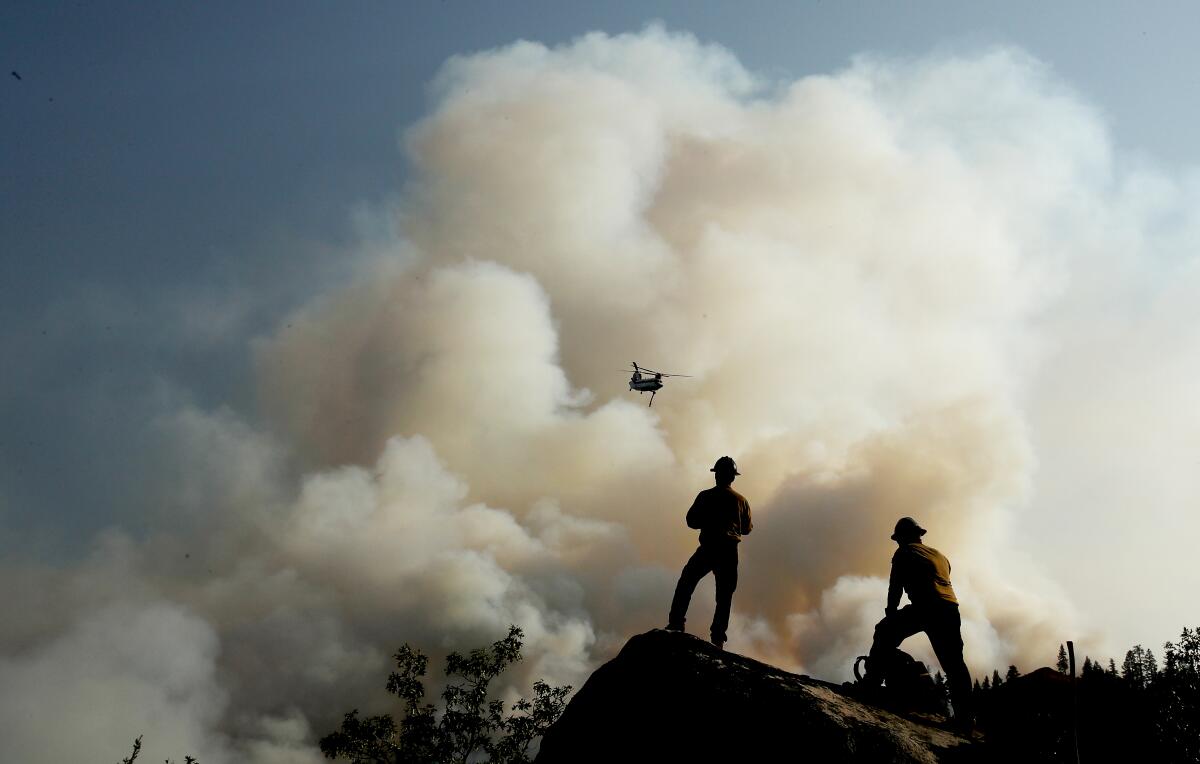 Two silhouetted firefighters watch a helicopter fly over a smoke cloud