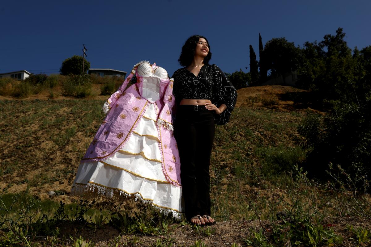 Karla Torres stands on a grassy area while holding an old-fashioned pink and white gown made from duct tape.