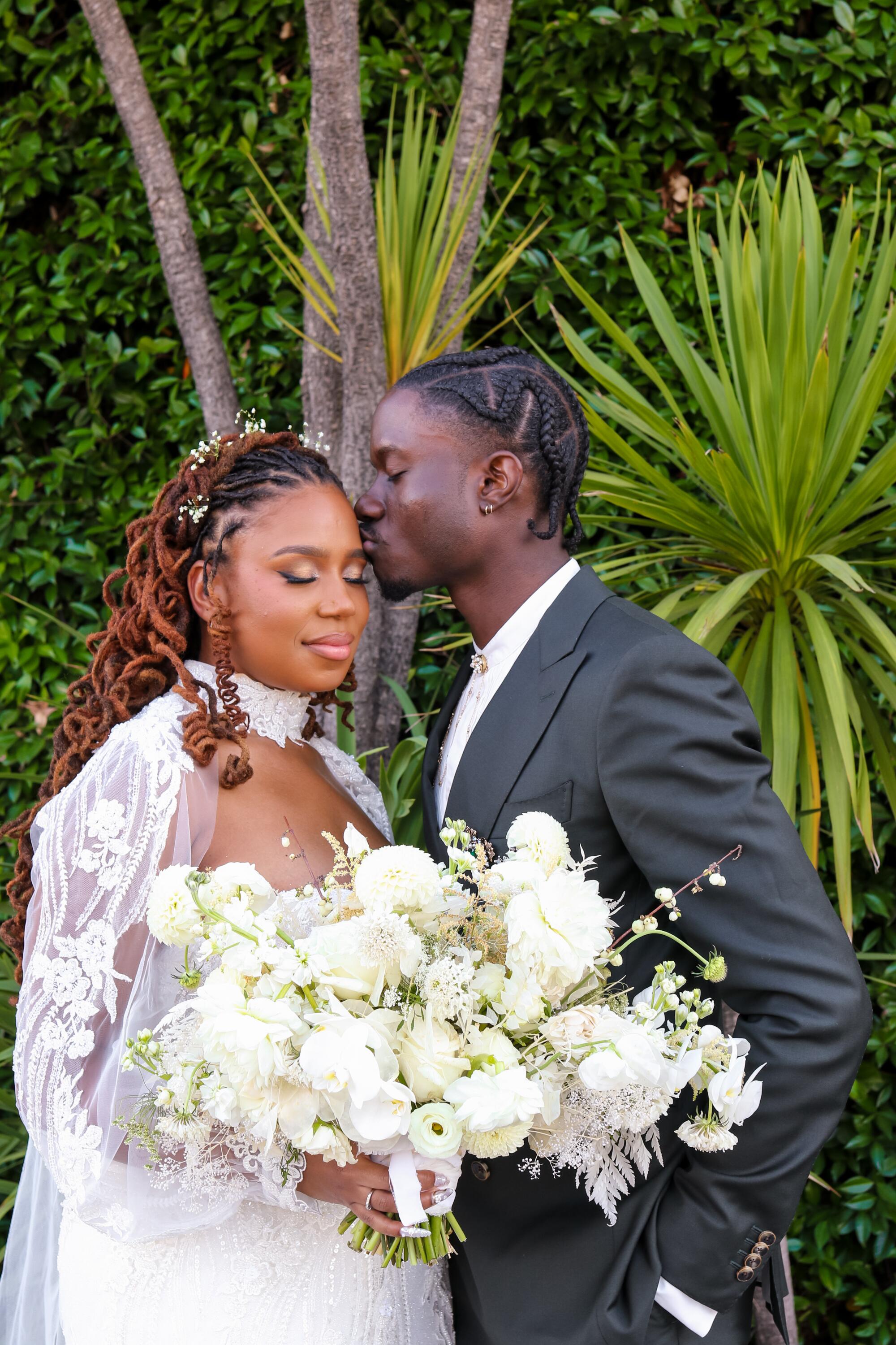 A man in a dark suit kisses his bride's forehead. She holds a big bouquet of white flowers.