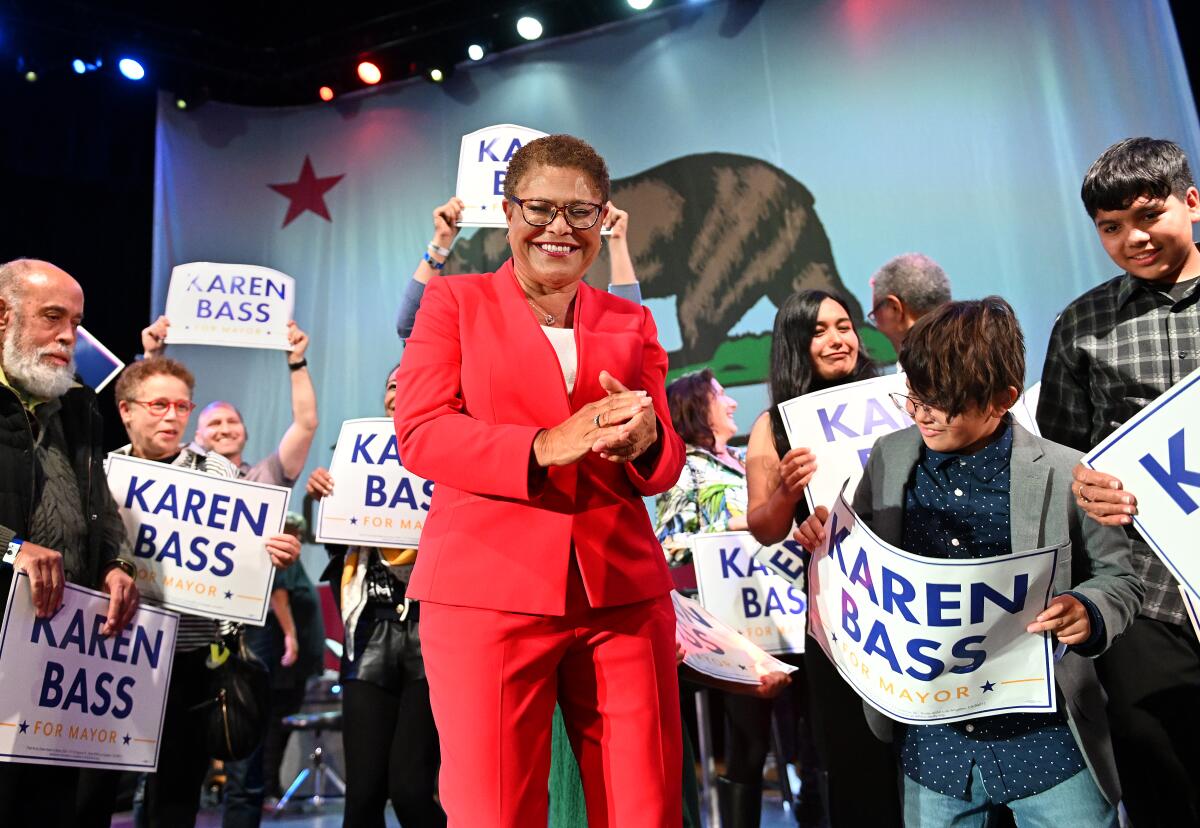 A woman in a red suit and glasses is seen smiling before supporters holding placards.