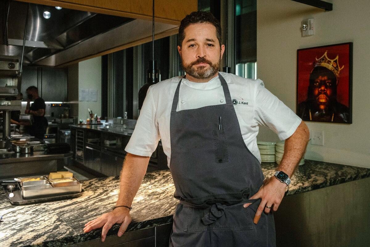 Chef James Kent, in a gray apron, stands at the bar of a restaurant with one hand on his hip.