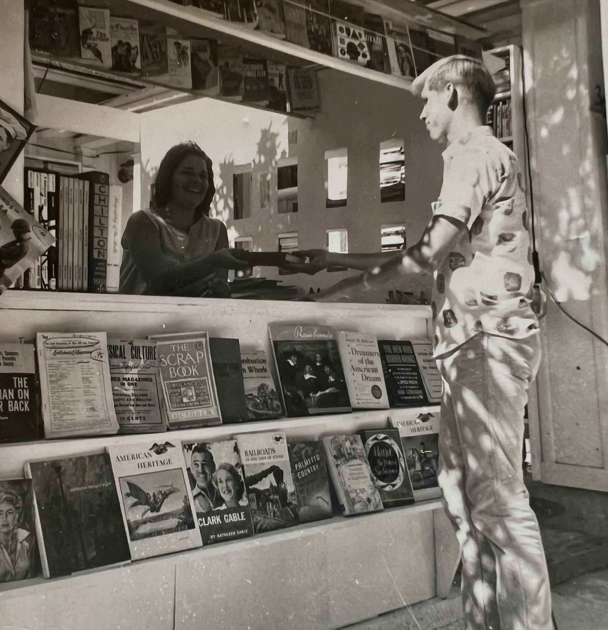 A black-and-white photo of a young woman behind a sales counter at Bart's, selling a book to a young man.