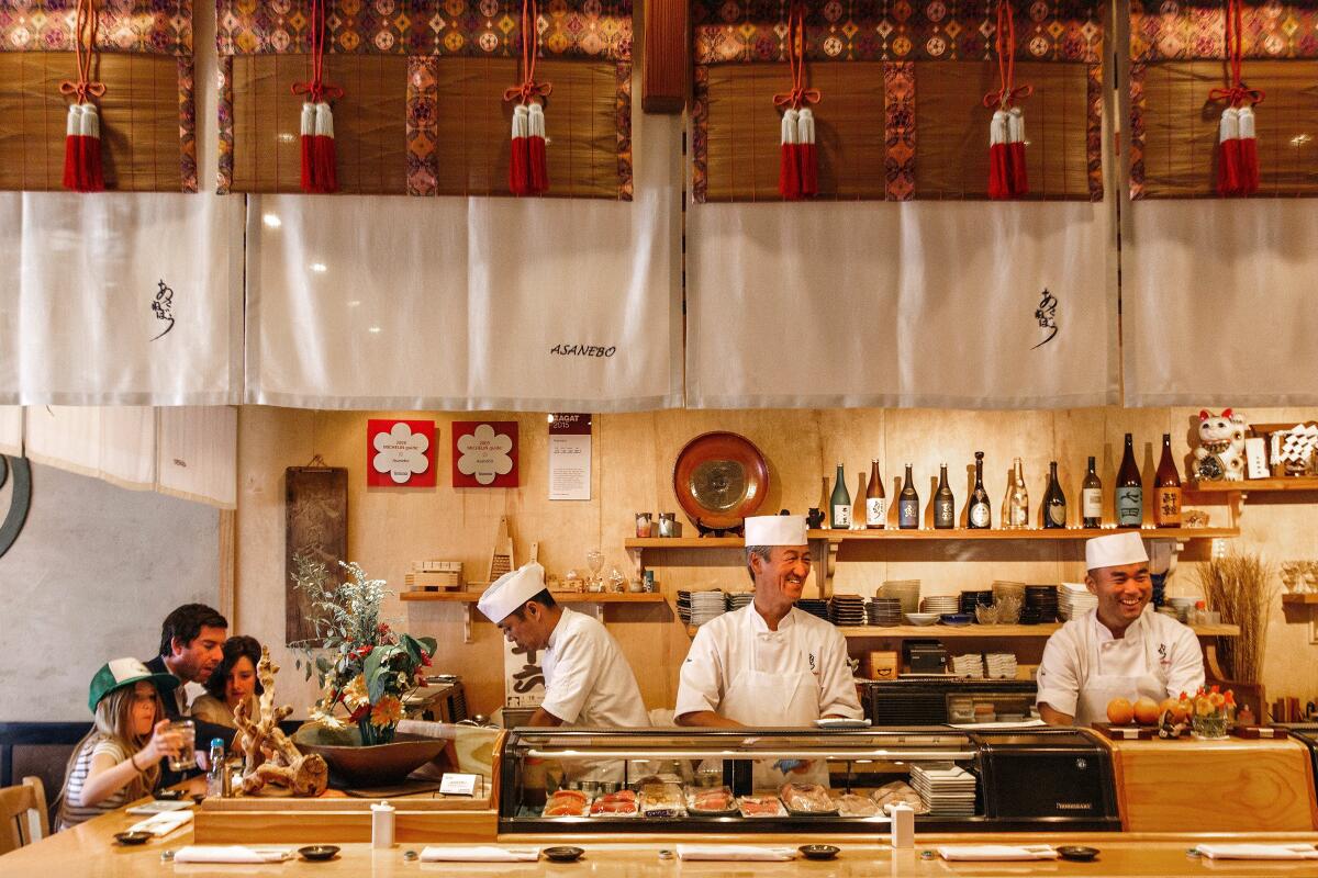 Three people at a sushi bar, with three smiling chefs standing behind the bar