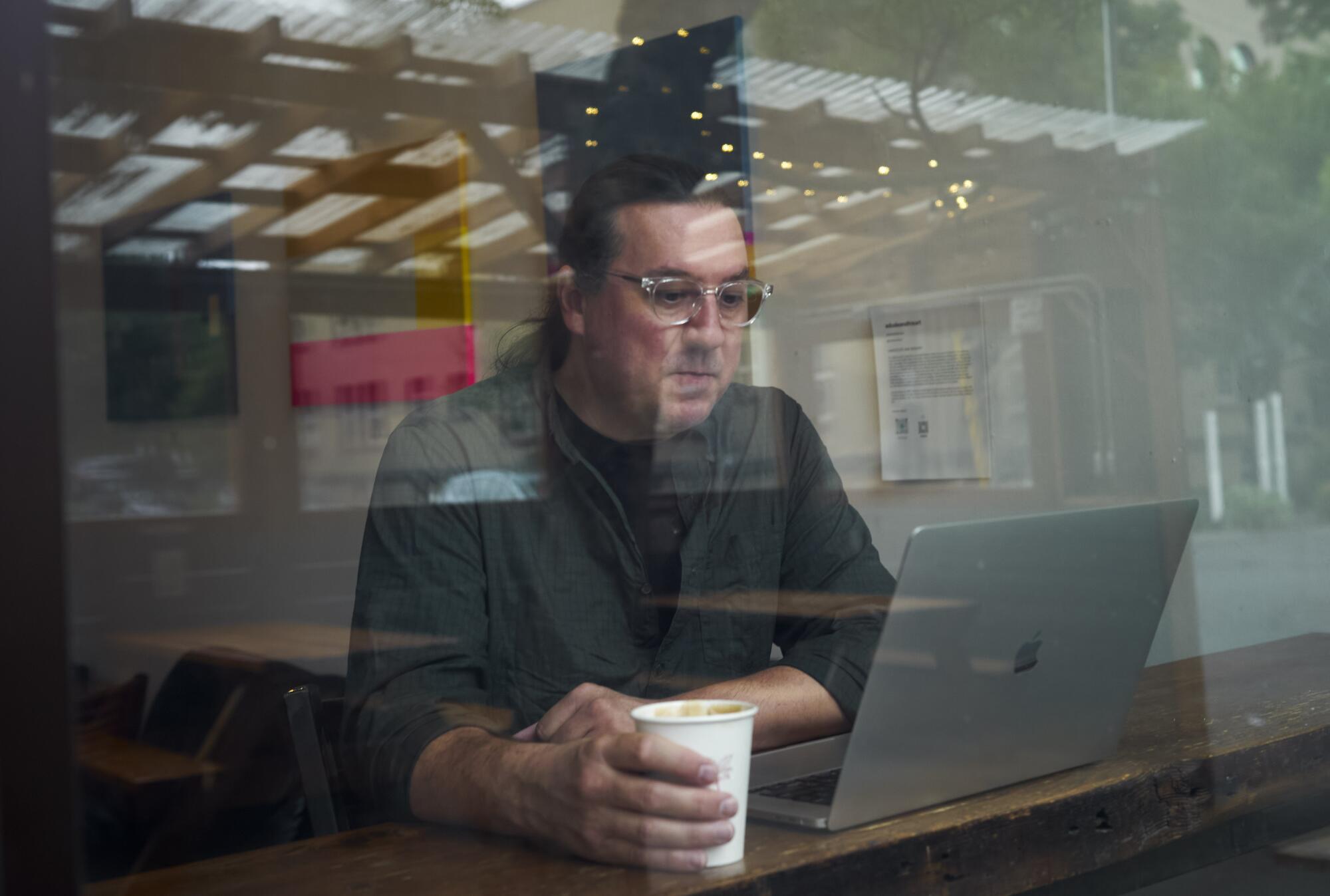 A man with glasses and a ponytail studies his computer screen at a coffee shop. 