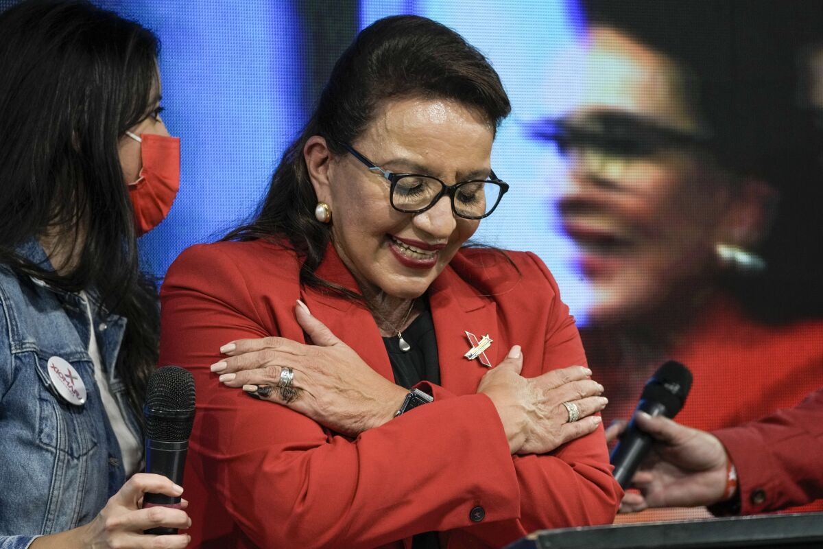 A woman with dark hair, wearing glasses and a red jacket, smiles as she looks down with her arms crossed