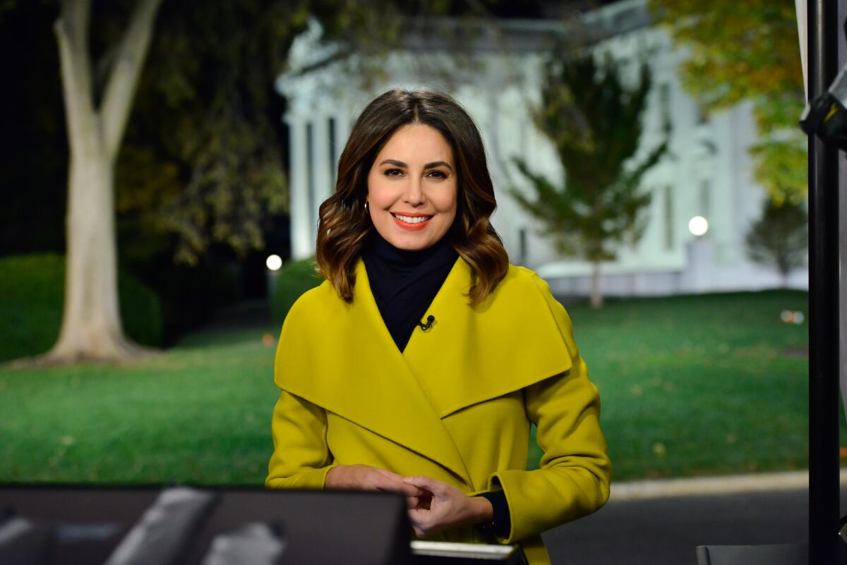 A woman wearing a chartreuse coat over a black turtleneck stands at night with the White House in the background
