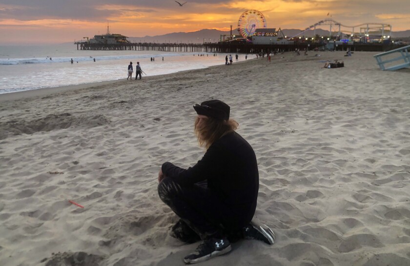 A man sits in the sand with the pier and beach in the background at sunset