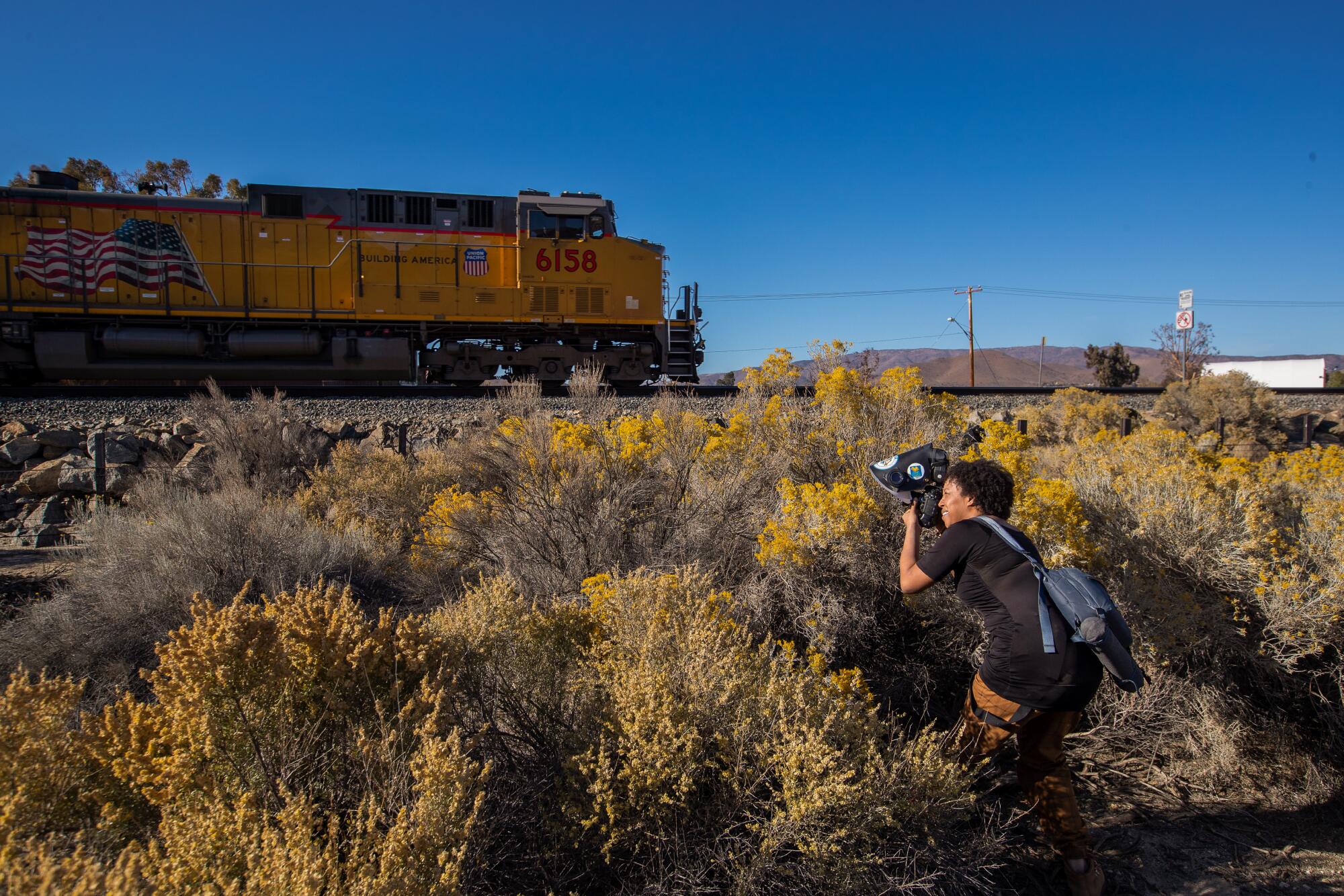 Krystle Hickman crouching to take pictures, with a train in the background of the desert scene.