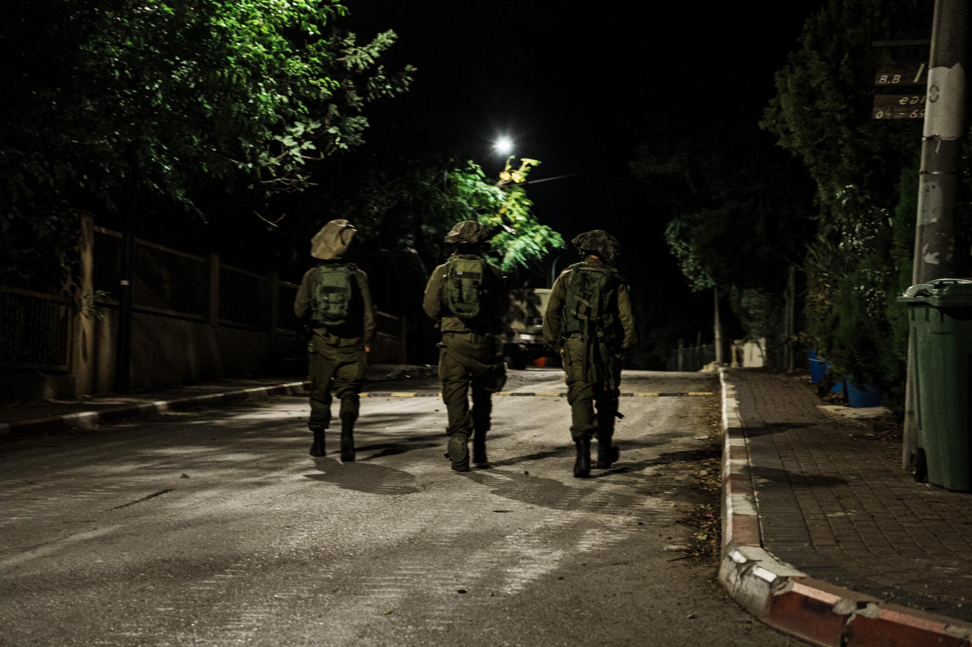 Three people in military uniform, seen from behind, walk on a darkened, empty street toward a light in the distance
