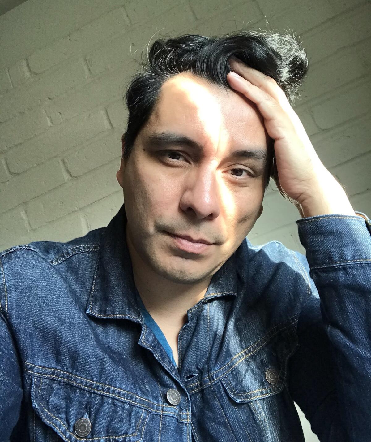 Manuel Muñoz, wearing a denim shirt, holds his head in his hand while seated before a white brick wall.