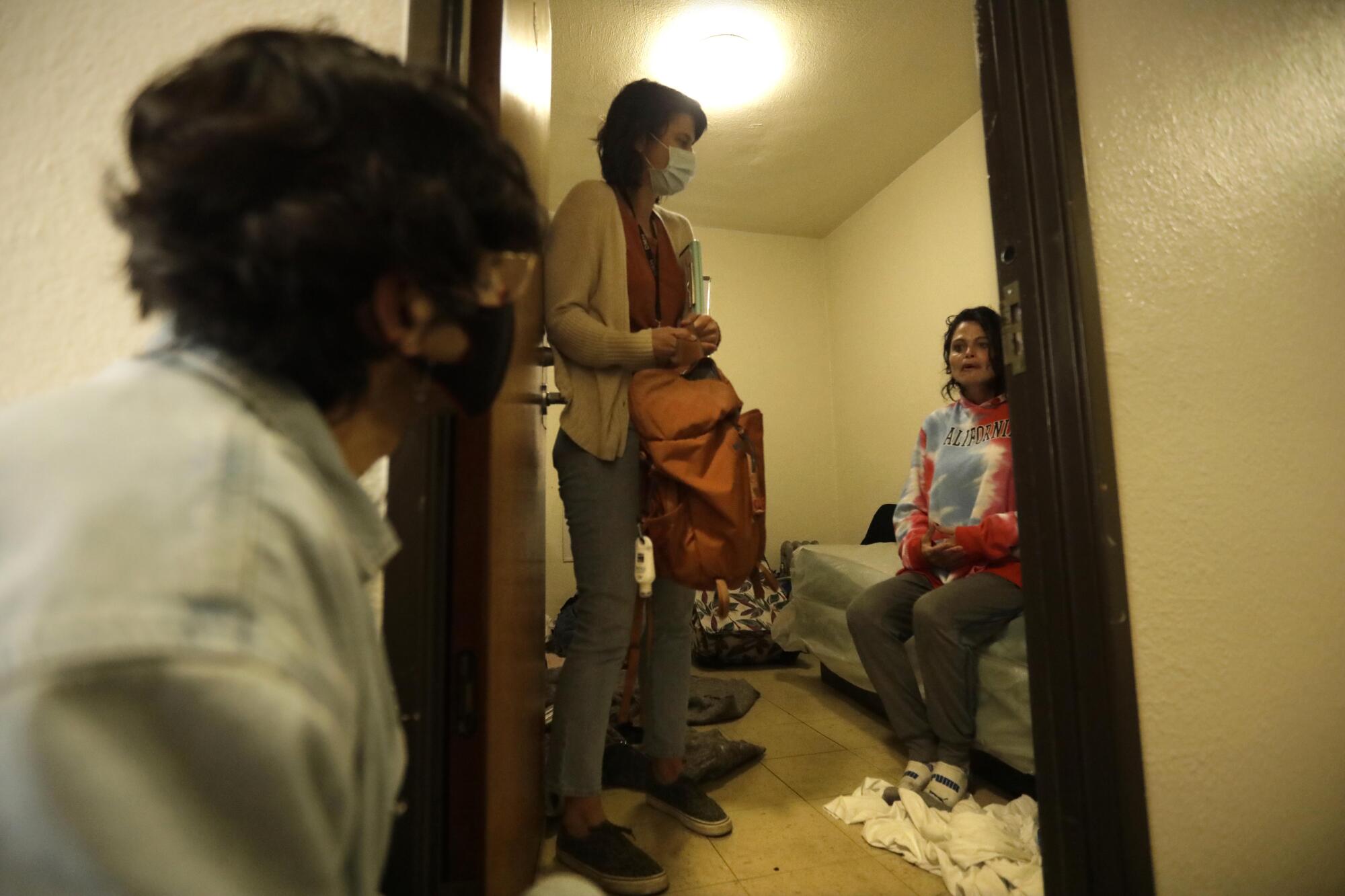 Two people look at a person sitting on a bed through a doorway.