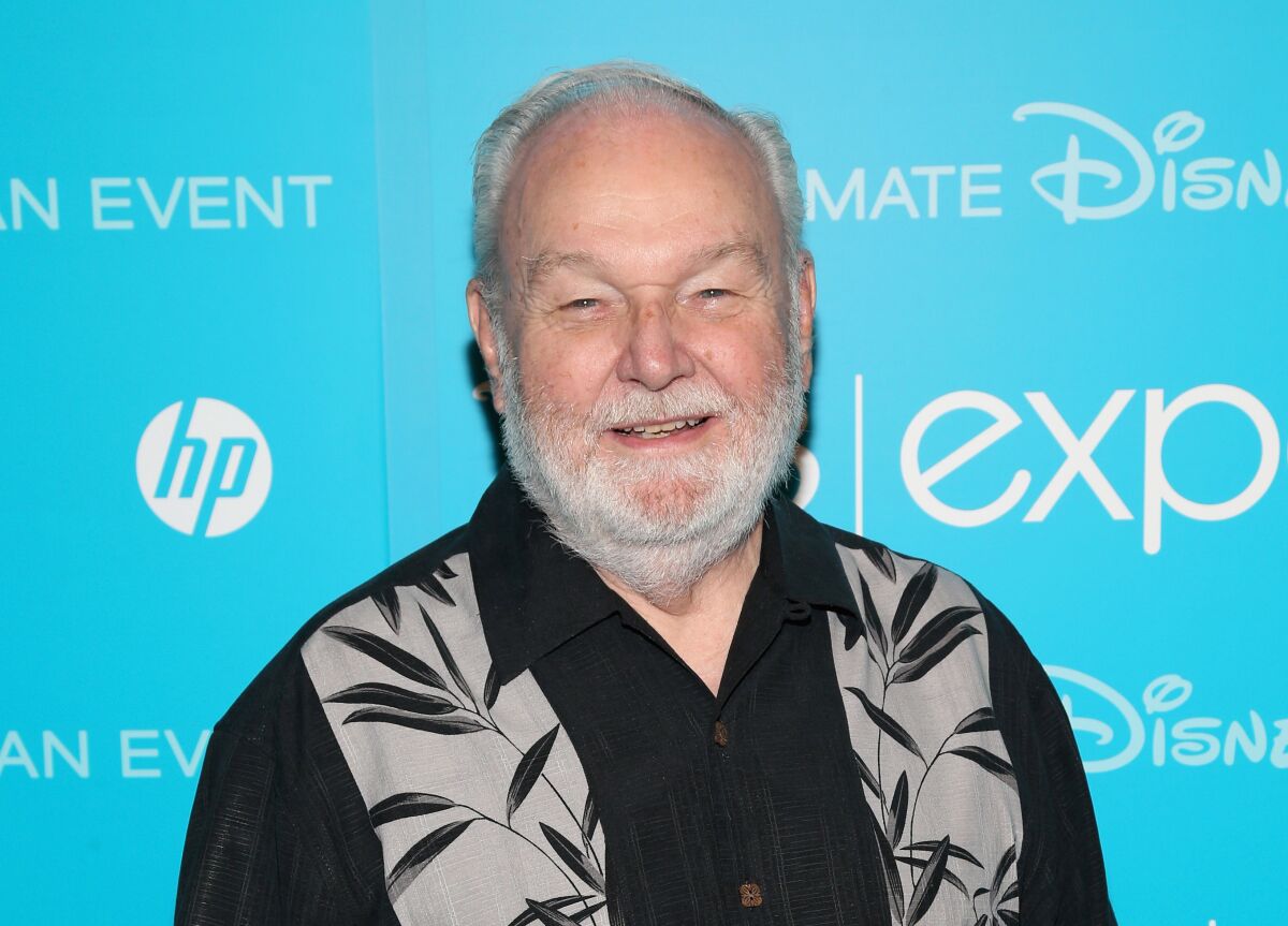 A man with gray hair and facial hair wearing a black shirt with a leaf print smiling