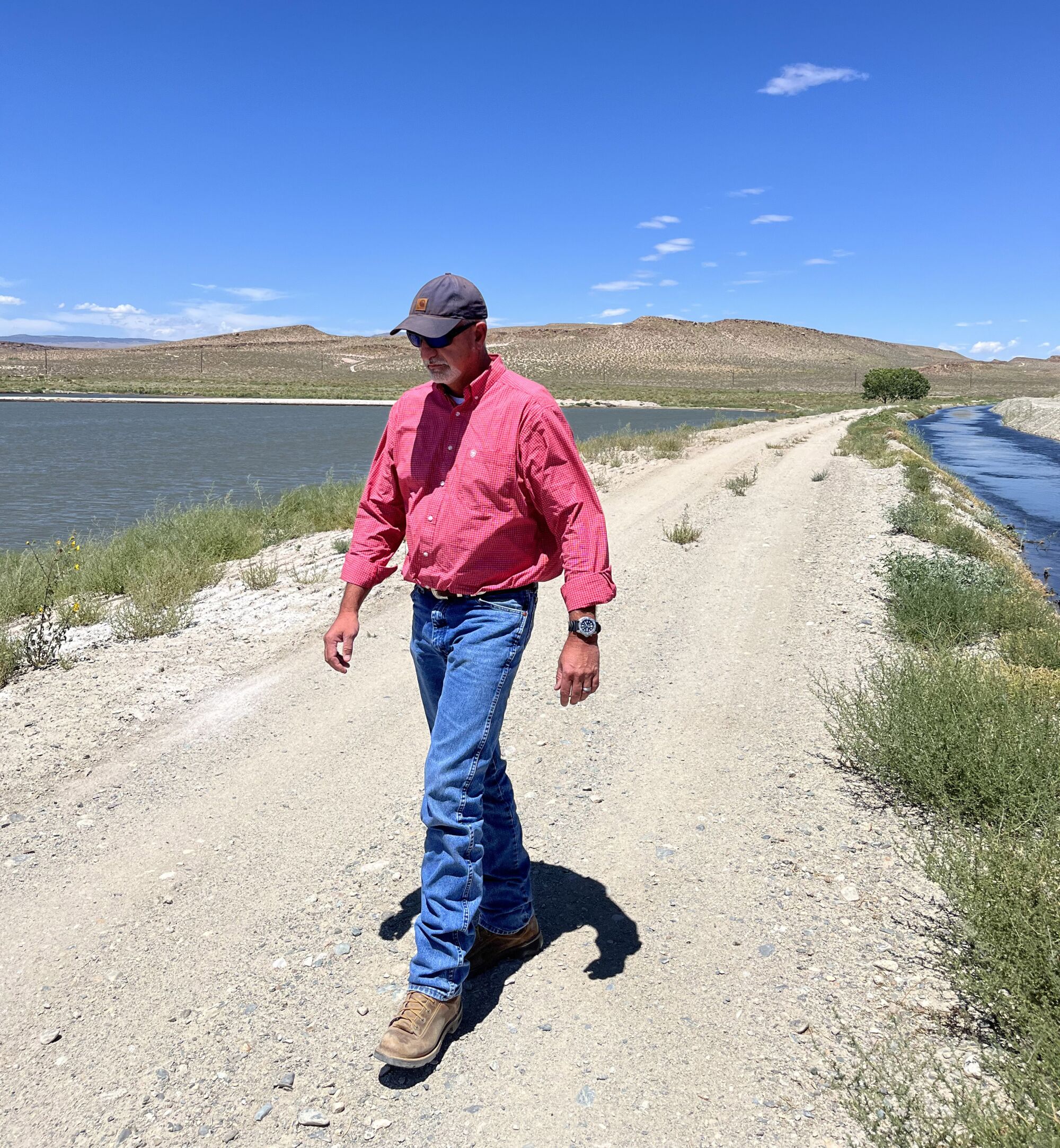 A man in a red shirt and blue jeans walks along a dirt road near a catch basin.