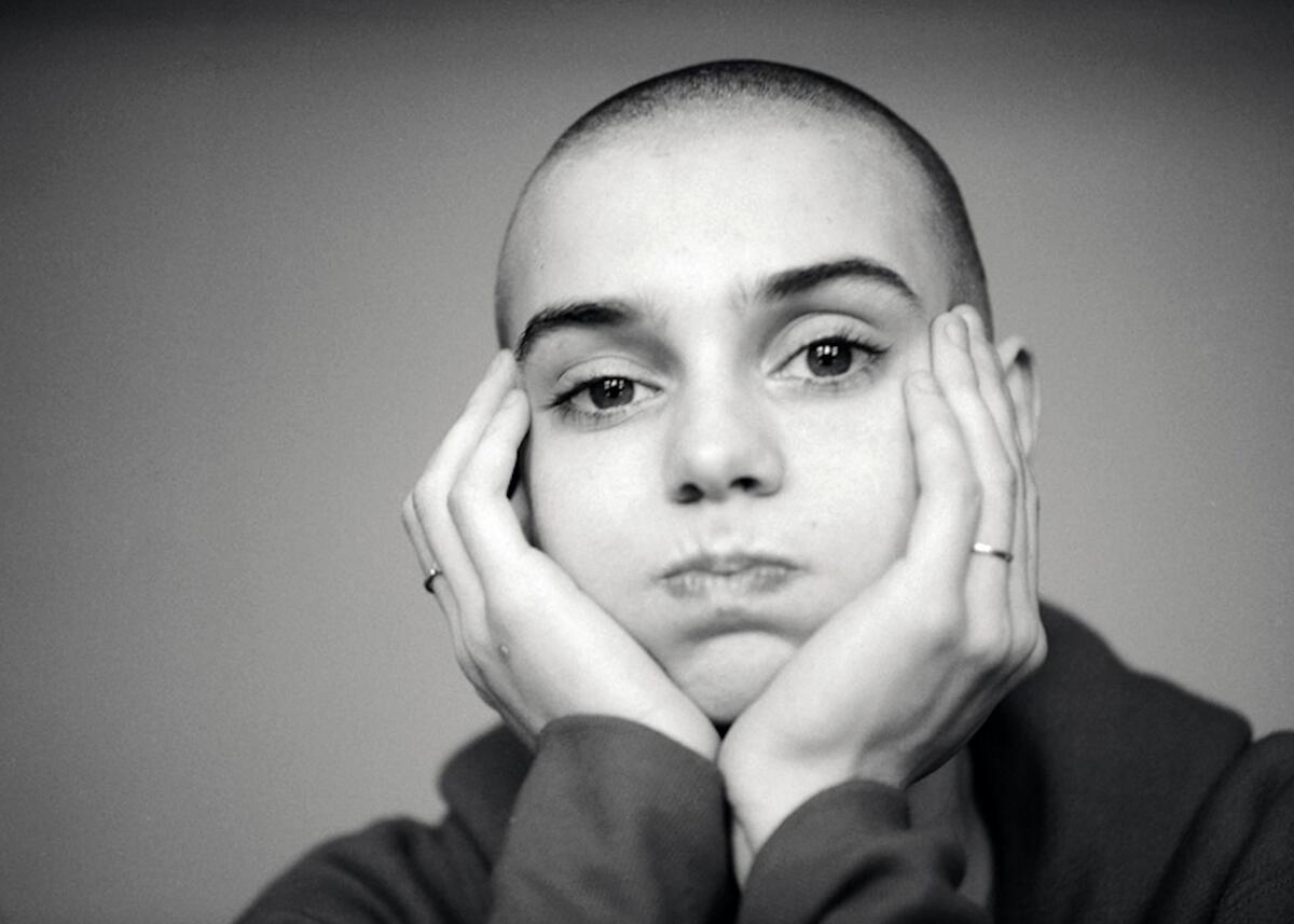 A black-and-white horizontal frame of Sinéad O'Connor resting her head in the palms of her hands, holding air in her cheeks
