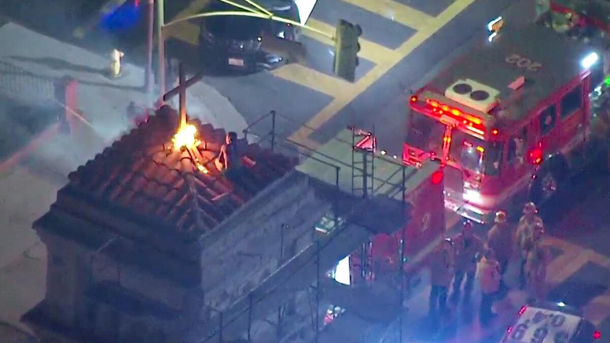 With Fire Department personnel standing below, a man crouches next to a fire atop a church.