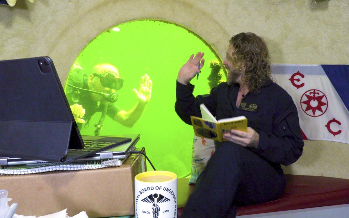 A man sits next to a porthole framing bright-green water and scuba diver swimming by