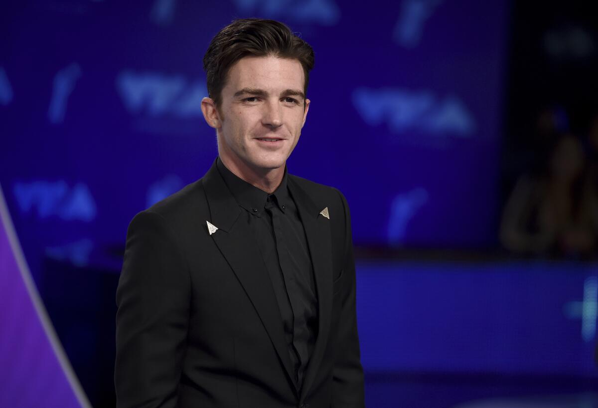 Drake Bell in a black shirt and suit standing against a blue backdrop.