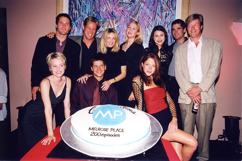 A group of 10 adults gathered around a cake.The cake is big, white and round and features a blue circle in the middle.