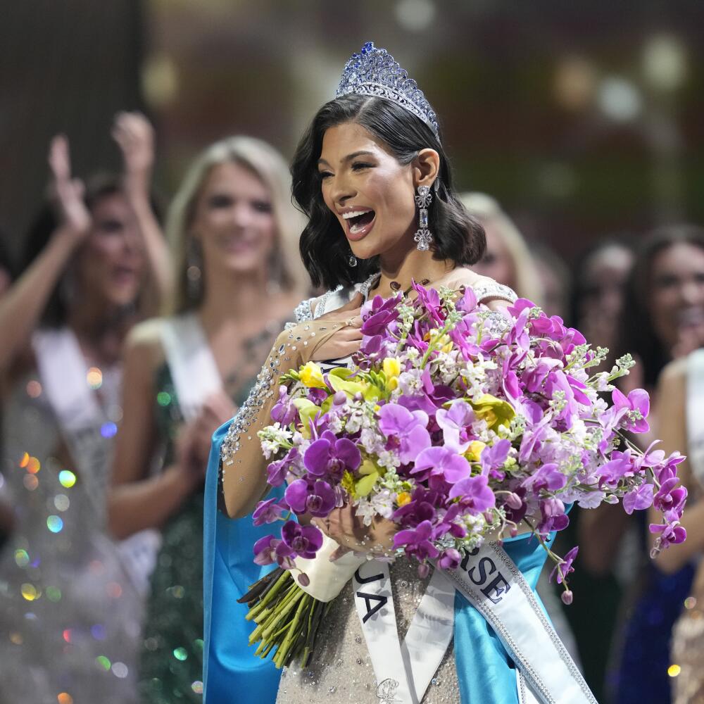 A woman with dark hair, wearing a crown, holds a large purple-and-yellow bouquet as other women applaud 