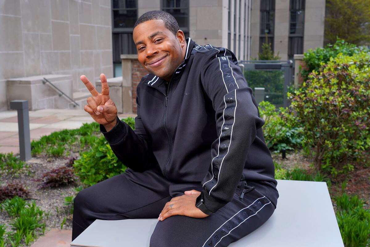 A smiling man wearing a tracksuit and sitting on a platform in a garden holds up two fingers in a peace sign.
